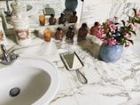A marble countertop in a bathroom with a white ceramic sink and silver faucet. On the counter are decorative ceramic canisters, some labeled, and a group of small amber-colored bottles. There is a handheld mirror with a metal handle, and a vase with pink flowers. The scene is reflected in a wall mirror behind the objects.