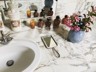 A marble countertop in a bathroom with a white ceramic sink and silver faucet. On the counter are decorative ceramic canisters, some labeled, and a group of small amber-colored bottles. There is a handheld mirror with a metal handle, and a vase with pink flowers. The scene is reflected in a wall mirror behind the objects.