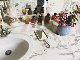 A marble countertop in a bathroom with a white ceramic sink and silver faucet. On the counter are decorative ceramic canisters, some labeled, and a group of small amber-colored bottles. There is a handheld mirror with a metal handle, and a vase with pink flowers. The scene is reflected in a wall mirror behind the objects.
