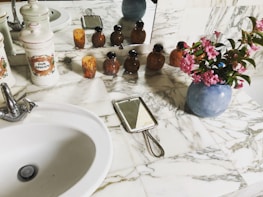A marble countertop in a bathroom with a white ceramic sink and silver faucet. On the counter are decorative ceramic canisters, some labeled, and a group of small amber-colored bottles. There is a handheld mirror with a metal handle, and a vase with pink flowers. The scene is reflected in a wall mirror behind the objects.