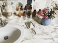 A marble countertop in a bathroom with a white ceramic sink and silver faucet. On the counter are decorative ceramic canisters, some labeled, and a group of small amber-colored bottles. There is a handheld mirror with a metal handle, and a vase with pink flowers. The scene is reflected in a wall mirror behind the objects.