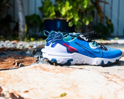 A brightly colored athletic shoe placed on a textured stone surface outdoors, surrounded by greenery in the background. The shoe features a predominantly blue upper with black laces, white sole with wavy patterns, and red accents.