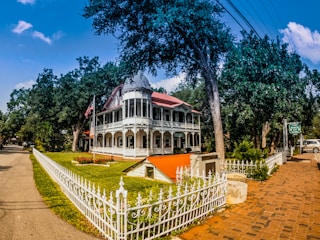 A large, ornate Victorian-style house with a wraparound porch and decorative white railings is surrounded by lush greenery and trees. An American flag is prominently displayed near the entrance, and a brick walkway leads towards the house. The architecture features a red roof and detailed woodwork.