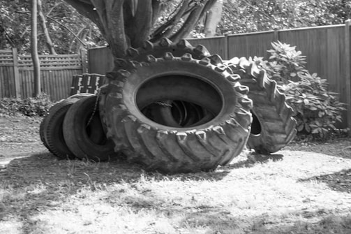 A group of large tractor tires is placed on a grassy area near a wooden fence. Some tires are stacked while others are leaning against a tree. The scene is outdoors, surrounded by foliage and trees nearby.