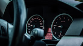 A close-up view of a car dashboard featuring two dials, including a speedometer and a tachometer. The speedometer displays speeds in kilometers per hour, and the tachometer measures engine RPMs. A digital screen shows additional information with a red backlight. The steering wheel is partially visible.