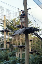 A person wearing shorts and a t-shirt stands on a wooden platform in a ropes course, high among trees. They are equipped with a safety harness and holding onto ropes attached to wooden posts. The background features a structure of nets and ropes among lush green foliage.