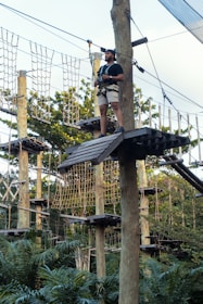 A person wearing shorts and a t-shirt stands on a wooden platform in a ropes course, high among trees. They are equipped with a safety harness and holding onto ropes attached to wooden posts. The background features a structure of nets and ropes among lush green foliage.