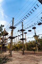 A ropes course with multiple wooden platforms and suspended bridges is set among tall poles. The structures are connected by various types of rope bridges and ladders, creating a challenging adventure course. Birds are flying in the clear blue sky above, and there are lush green trees surrounding the area, providing a natural backdrop. Individuals are seen navigating the high ropes, indicating an outdoor recreational activity.