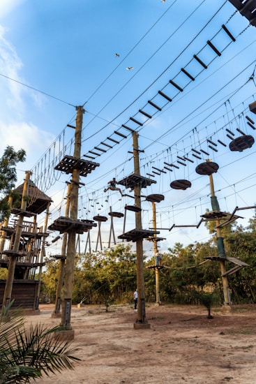 A ropes course with multiple wooden platforms and suspended bridges is set among tall poles. The structures are connected by various types of rope bridges and ladders, creating a challenging adventure course. Birds are flying in the clear blue sky above, and there are lush green trees surrounding the area, providing a natural backdrop. Individuals are seen navigating the high ropes, indicating an outdoor recreational activity.