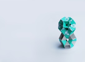 A geometric sculpture composed of interlocking paper shapes, primarily using teal and grey colors, stands against a minimalist light grey background. The shapes are arranged in a compact, stacked formation.