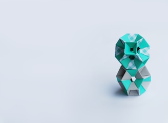 A geometric sculpture composed of interlocking paper shapes, primarily using teal and grey colors, stands against a minimalist light grey background. The shapes are arranged in a compact, stacked formation.