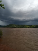 Dark, heavy clouds gather over a wide river, hinting at an approaching storm. The water appears brownish and reflects the gloomy sky. Lush green hills line the opposite bank, adding contrast to the ominous atmosphere. A few trees are visible on the edges, framing the scene.