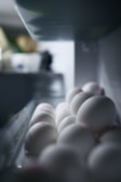 A close-up view of white eggs neatly arranged in a refrigerator tray. The eggs appear smooth and have faint pink markings on top. The lighting casts soft shadows, creating a serene and clean atmosphere.