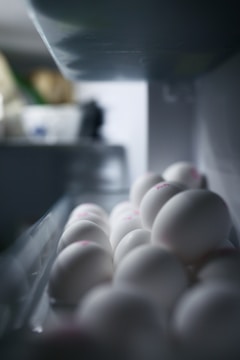A close-up view of white eggs neatly arranged in a refrigerator tray. The eggs appear smooth and have faint pink markings on top. The lighting casts soft shadows, creating a serene and clean atmosphere.