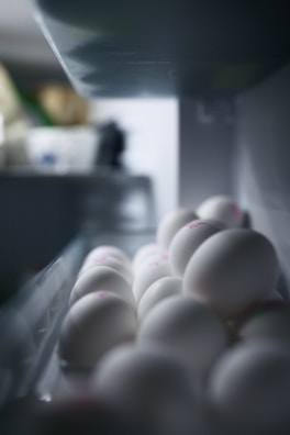 A close-up view of white eggs neatly arranged in a refrigerator tray. The eggs appear smooth and have faint pink markings on top. The lighting casts soft shadows, creating a serene and clean atmosphere.