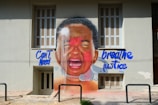 A large mural painted on the wall of a building depicts a young crying child with their face partially covered in red paint. The words 'Can't breathe' and 'Need justice' are written in large blue letters around the image. It is a powerful and emotive piece of street art.