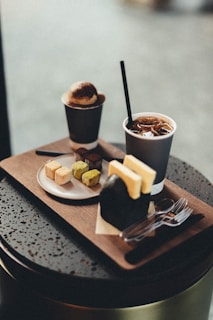 A wooden tray on a table featuring a variety of refreshments, including a cup of iced coffee with a straw, a smaller cup with a dessert topped with cocoa powder, a plate with cubed pastries in different flavors, and a wrapped sandwich or pastry with toast. Silverware is placed on the tray, adjacent to the food items.