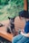 A woman wearing a mask is sitting on a wooden platform with a small, brindle dog sitting beside her. The dog looks towards the camera with its tongue out, appearing happy. The background is blurred with greenery, suggesting an outdoor setting.