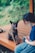 A woman wearing a mask is sitting on a wooden platform with a small, brindle dog sitting beside her. The dog looks towards the camera with its tongue out, appearing happy. The background is blurred with greenery, suggesting an outdoor setting.