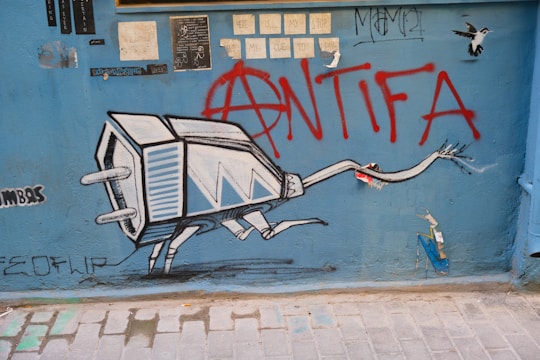 A vibrant piece of street art features a robotic insect with a television or plug head, set against a blue wall with various stickers and smaller graffiti pieces. Prominently, the word 'ANTIFA' and an anarchy symbol are spray-painted in red over the artwork. Additional smaller details include different tags, a bird, and some handwritten notes.