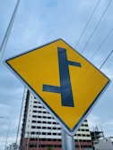 A yellow road sign with a black T-intersection symbol is prominently displayed. In the background, there is a partially constructed building with scaffolding, and gray skies overhead. Power lines cross horizontally above the sign, giving an urban feel.