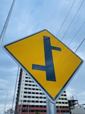 A yellow road sign with a black T-intersection symbol is prominently displayed. In the background, there is a partially constructed building with scaffolding, and gray skies overhead. Power lines cross horizontally above the sign, giving an urban feel.