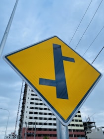 A yellow road sign with a black T-intersection symbol is prominently displayed. In the background, there is a partially constructed building with scaffolding, and gray skies overhead. Power lines cross horizontally above the sign, giving an urban feel.
