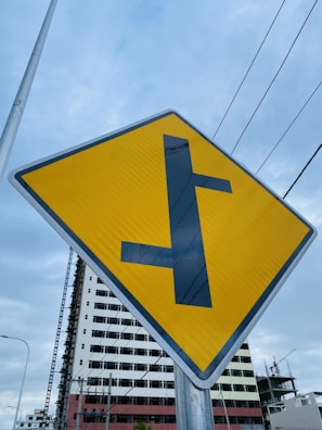 A yellow road sign with a black T-intersection symbol is prominently displayed. In the background, there is a partially constructed building with scaffolding, and gray skies overhead. Power lines cross horizontally above the sign, giving an urban feel.