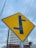 A yellow road sign with a black T-intersection symbol is prominently displayed. In the background, there is a partially constructed building with scaffolding, and gray skies overhead. Power lines cross horizontally above the sign, giving an urban feel.