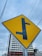 A yellow road sign with a black T-intersection symbol is prominently displayed. In the background, there is a partially constructed building with scaffolding, and gray skies overhead. Power lines cross horizontally above the sign, giving an urban feel.