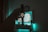 A smartwatch is held up against a blurred background of a room illuminated by teal lighting. The watch face displays the time with large, bold numerals. The setting includes a computer screen that emits soft, colorful light, contributing to a calm and modern atmosphere.