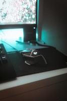 A computer setup featuring a mechanical gaming mouse with illuminated accents on a black mouse pad. The mouse is positioned next to a keyboard, and a monitor displays a blurry, colorful background. Ambient teal lighting creates a futuristic and gaming-themed atmosphere.