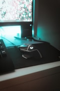 A computer setup featuring a mechanical gaming mouse with illuminated accents on a black mouse pad. The mouse is positioned next to a keyboard, and a monitor displays a blurry, colorful background. Ambient teal lighting creates a futuristic and gaming-themed atmosphere.