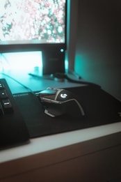 A computer setup featuring a mechanical gaming mouse with illuminated accents on a black mouse pad. The mouse is positioned next to a keyboard, and a monitor displays a blurry, colorful background. Ambient teal lighting creates a futuristic and gaming-themed atmosphere.