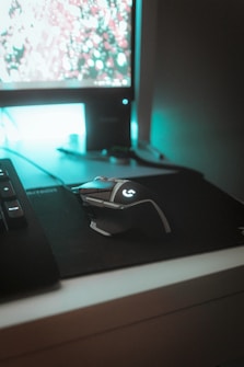 A computer setup featuring a mechanical gaming mouse with illuminated accents on a black mouse pad. The mouse is positioned next to a keyboard, and a monitor displays a blurry, colorful background. Ambient teal lighting creates a futuristic and gaming-themed atmosphere.