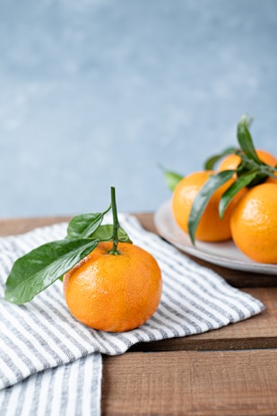 Fresh, ripe oranges sit on a wooden table. One orange with a green leaf rests on a neatly folded striped cloth in the foreground, while more oranges on a plate are visible in the background. The setting has a rustic and natural feel.