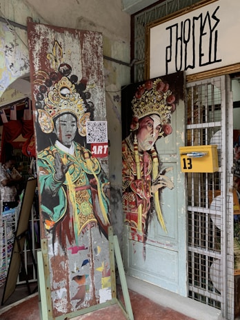 The image features two artworks depicting figures in traditional ornate costumes, possibly representing theatrical characters. The paintings are mounted on old, weathered boards leaning against a shop entrance with a yellow mailbox labeled '13'. A QR code is visible on one artwork, and there is an unoccupied metal display grid and an open door partially covered by the art.