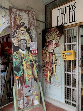 The image features two artworks depicting figures in traditional ornate costumes, possibly representing theatrical characters. The paintings are mounted on old, weathered boards leaning against a shop entrance with a yellow mailbox labeled '13'. A QR code is visible on one artwork, and there is an unoccupied metal display grid and an open door partially covered by the art.