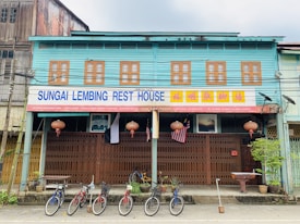 A two-story building with sea-green and brown wooden walls featuring a row of evenly spaced windows. A sign that reads 'Sungai Lembing Rest House' hangs above the entrance. Bicycles are parked along the sidewalk, and several traditional red lanterns and a flag are displayed. The building has a rustic, old-style architecture, reminiscent of traditional shop houses.