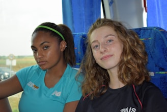 Two young women are seated inside a vehicle with blue upholstery. One has long brown hair tied with a green headband, wearing a light blue sports shirt, and the other has curly, light brown hair and is wearing a black shirt. The background shows a blurred outdoor landscape visible through a window.