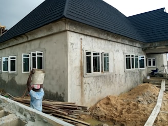 A house with a dark, sloped roof and multiple white-framed windows is under construction. The walls are unfinished, showing raw plasterwork, and raw materials like sand and planks are scattered around. A person is carrying a bucket on their head, walking along a narrow path next to the building.