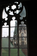 A gothic-style window with intricate designs is set in a dimly lit interior space. Through the window, an architectural structure with detailed stonework and a pointed tower is visible. The scene outside includes rooftops with greenish tones and a cloudy sky.