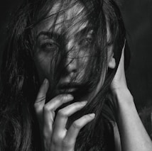 A black and white portrait of a person with long hair partially covering their face. The person's hand is positioned delicately near their mouth, adding an element of mystery and introspection. Their eyes gaze directly at the viewer, creating an intimate connection.