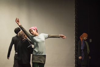 Several people are on stage, possibly rehearsing or performing. One person is in the foreground, extending an arm with a graceful gesture, wearing a headscarf and casual attire. Others in the background appear to be observing or participating in the activity. The setting has a minimal, focused ambiance.