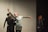 Several people are on stage, possibly rehearsing or performing. One person is in the foreground, extending an arm with a graceful gesture, wearing a headscarf and casual attire. Others in the background appear to be observing or participating in the activity. The setting has a minimal, focused ambiance.
