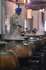 A person wearing a blue turban is stirring food in large metal pots, with steam rising from the pots in what appears to be an industrial kitchen setting.