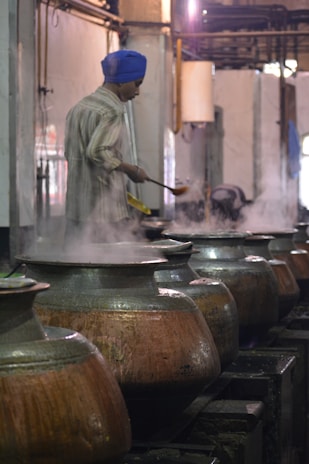 A person wearing a blue turban is stirring food in large metal pots, with steam rising from the pots in what appears to be an industrial kitchen setting.