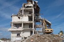 A partially demolished building with exposed beams, debris surrounding the base, and an excavator actively working on the site. The structure shows severe damage with torn walls and collapsed sections. The sky is clear and blue.