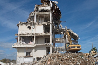 A partially demolished building with exposed beams, debris surrounding the base, and an excavator actively working on the site. The structure shows severe damage with torn walls and collapsed sections. The sky is clear and blue.