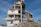 A partially demolished building with exposed beams, debris surrounding the base, and an excavator actively working on the site. The structure shows severe damage with torn walls and collapsed sections. The sky is clear and blue.